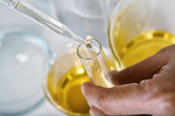 Hand hält ein Reagenzglas, in das mit einer Pipette eine goldgelbe Flüssigkeit geträufelt wird – im Hintergrund mehrere Glasgefäße mit derselben Substanz.